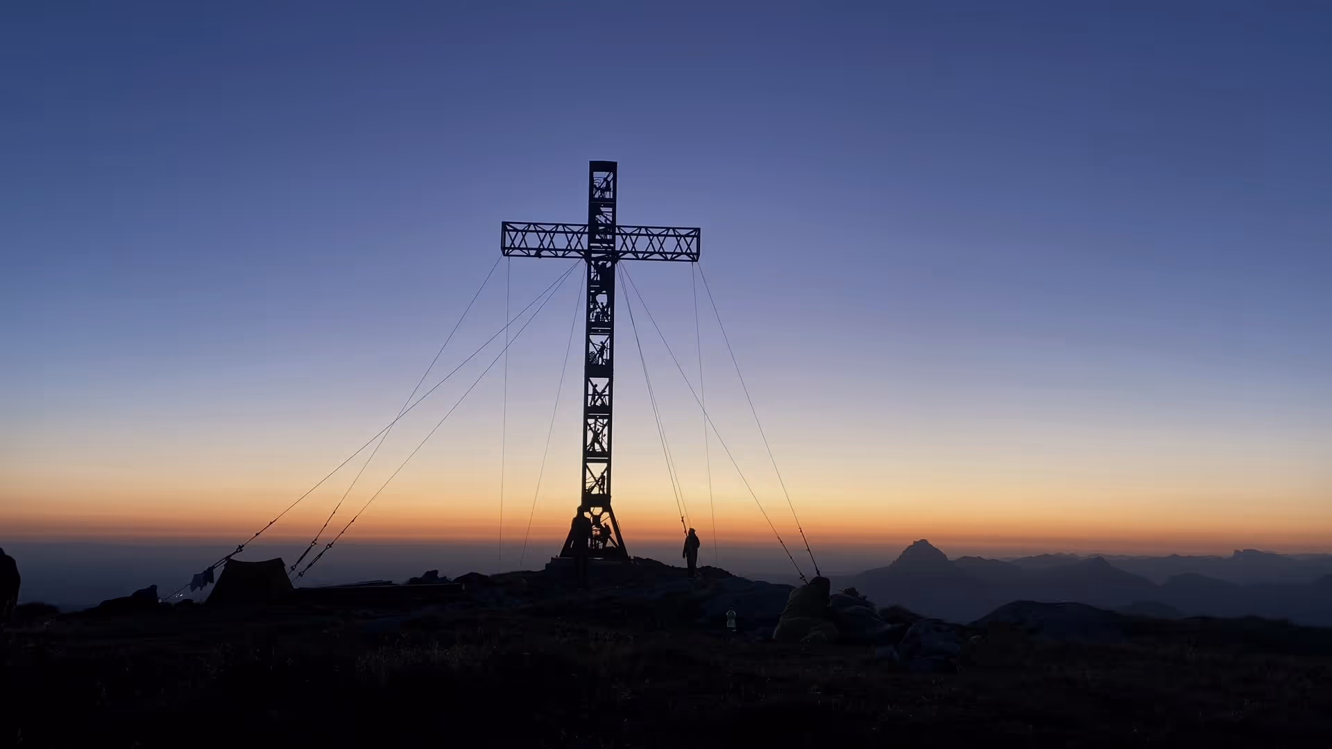 La cruz de cumbre de celosía en el Grünalmkogel recortada contra el amanecer, delante una tienda, sacos de dormir y dos siluetas