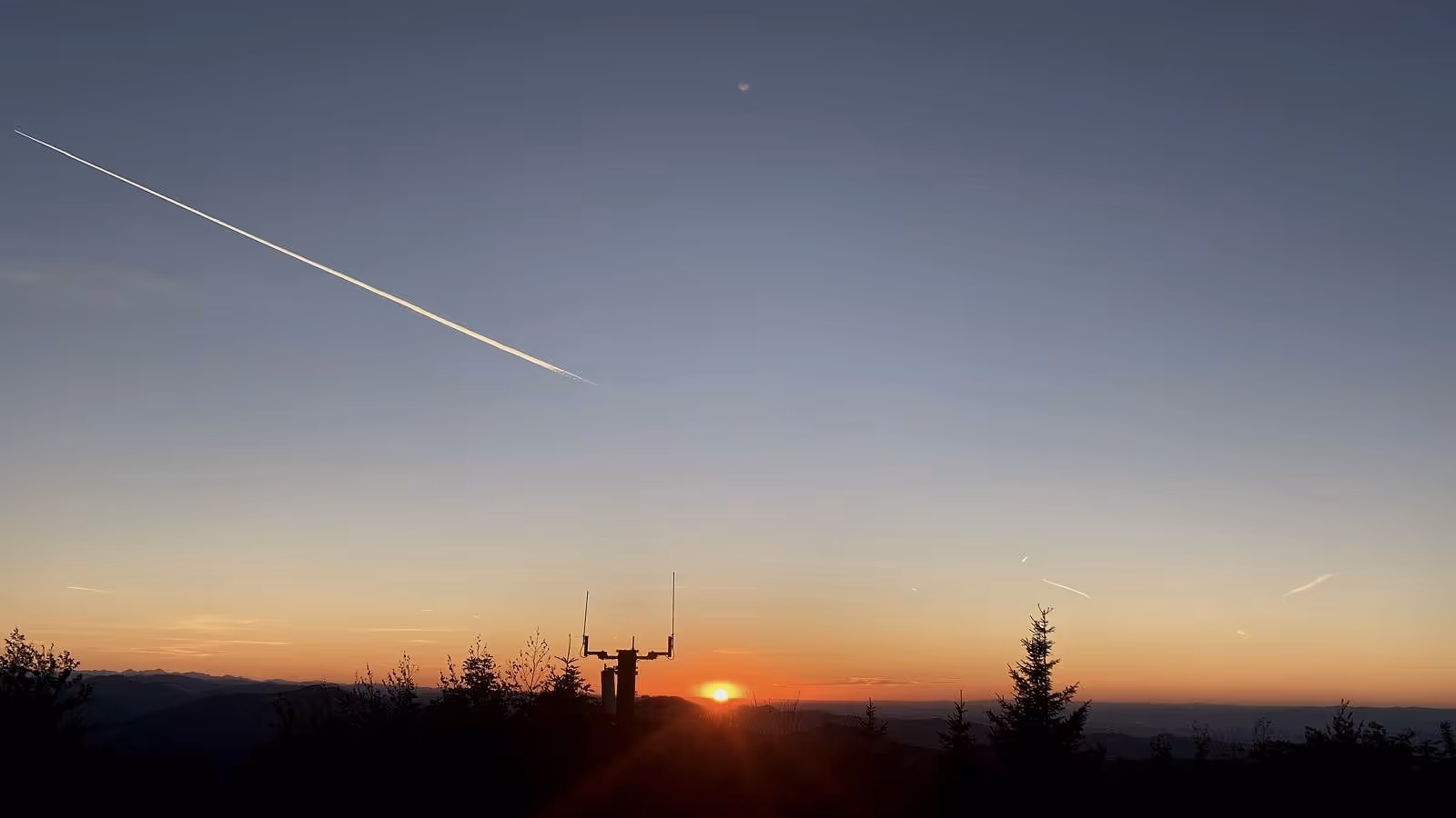 Un mástil de antena en silueta contra el cielo del crepúsculo, el sol tocando el horizonte, una estela encima