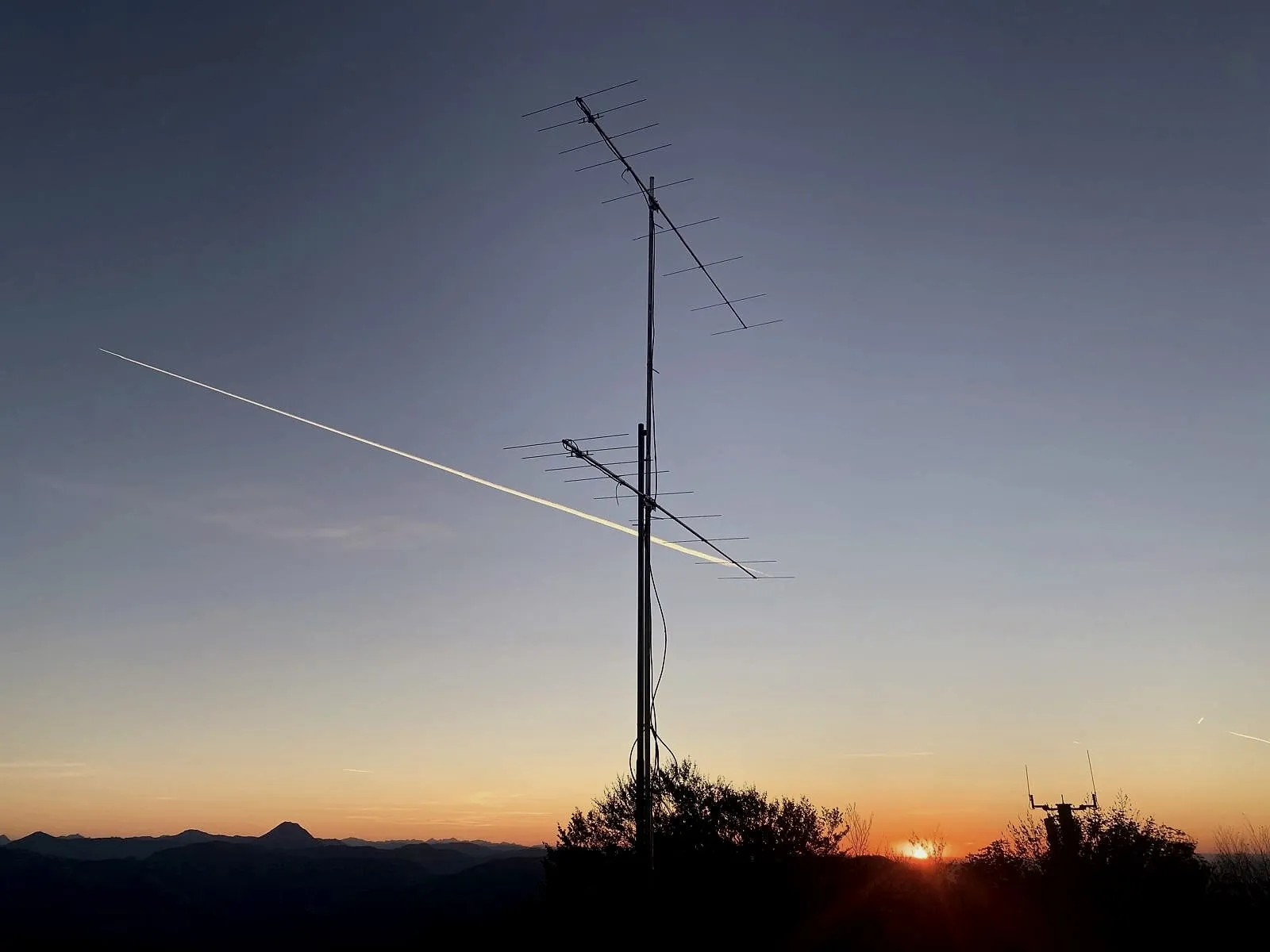 Dos yagis apiladas en el mástil contra el cielo de la tarde, una estela cruza toda la imagen, abajo las cadenas montañosas