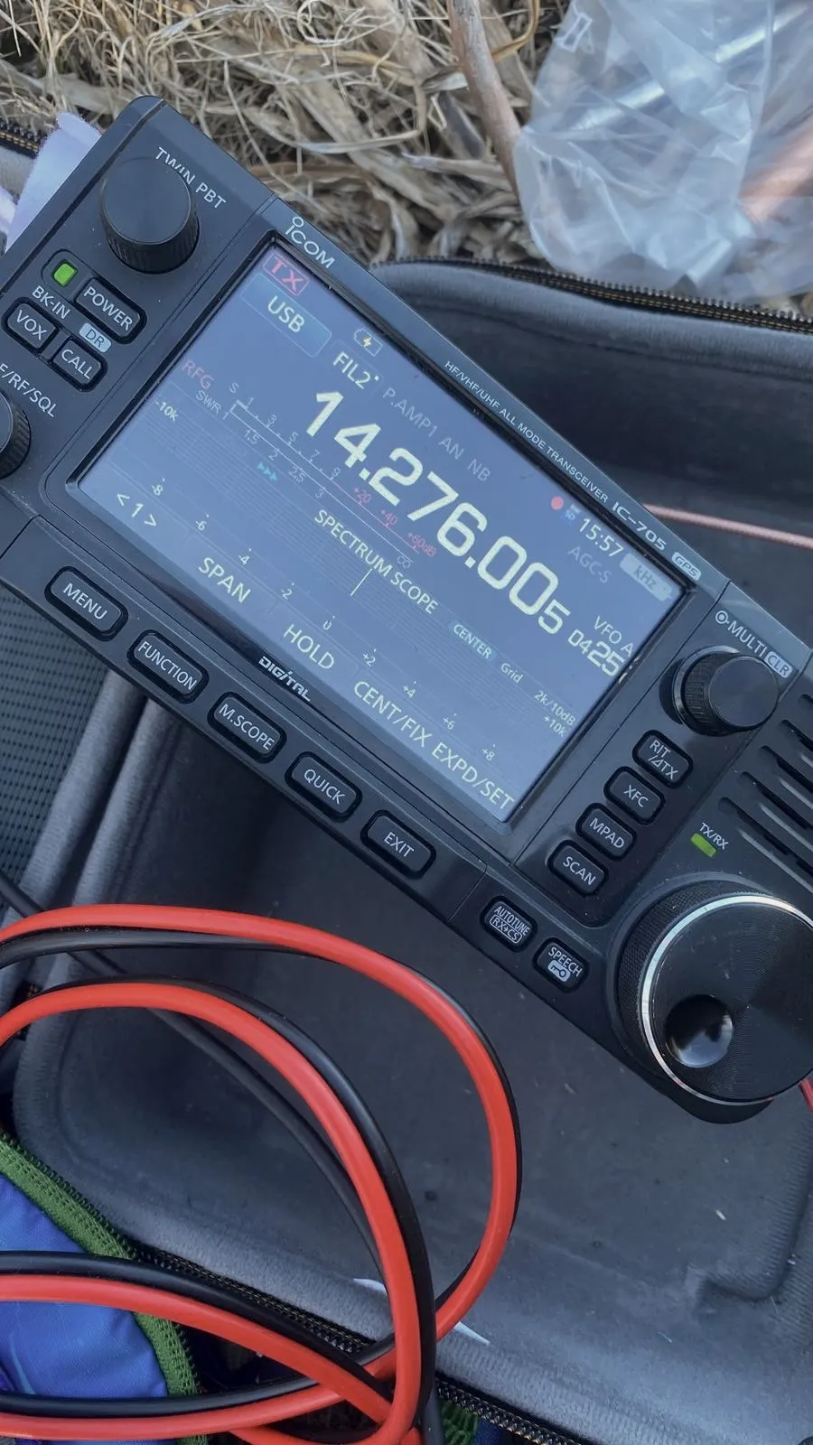 El Icom IC-705 apoyado sobre su bolsa, en la pantalla 14.276.005 en USB, al lado el cable de alimentación rojo y negro
